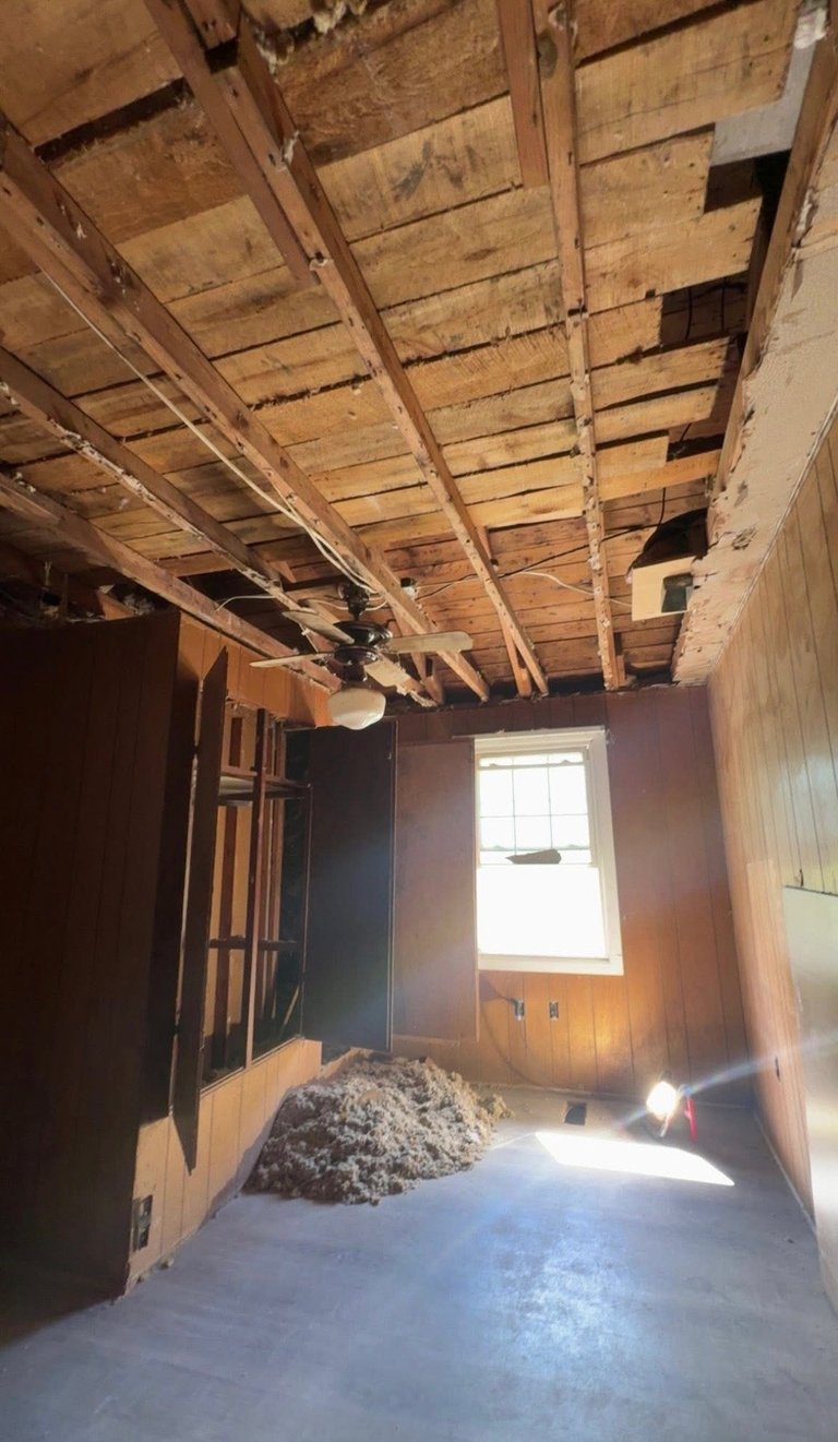 Interior of a room under renovation with exposed wooden ceiling joists, white walls, window with bright light, and debris pile on floor