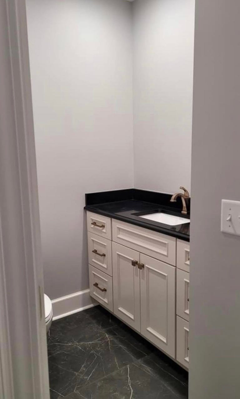 Small bathroom vanity with white cabinetry, black countertop, and integrated sink in a narrow white-walled space with dark marble flooring