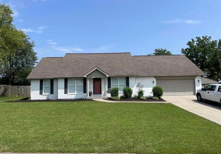 Single-story white and gray ranch-style home with brown roof, red door, driveway, and manicured lawn