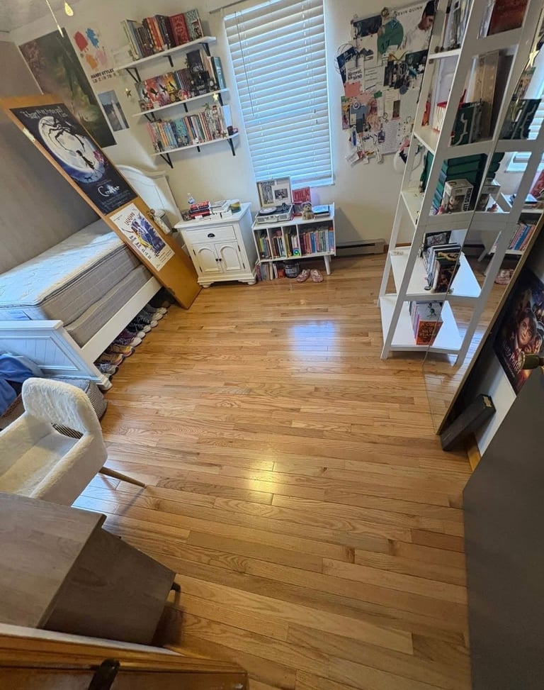 Overhead view of a bedroom or studio apartment with wooden floors, shelving units, posters, a window with blinds, and various storage furniture arranged throughout the space