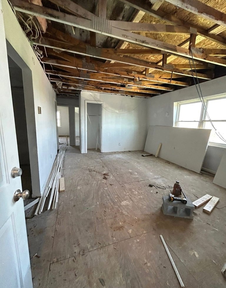 Interior of unfinished room under construction with exposed wooden ceiling joists, white drywall, concrete subfloor, and building materials