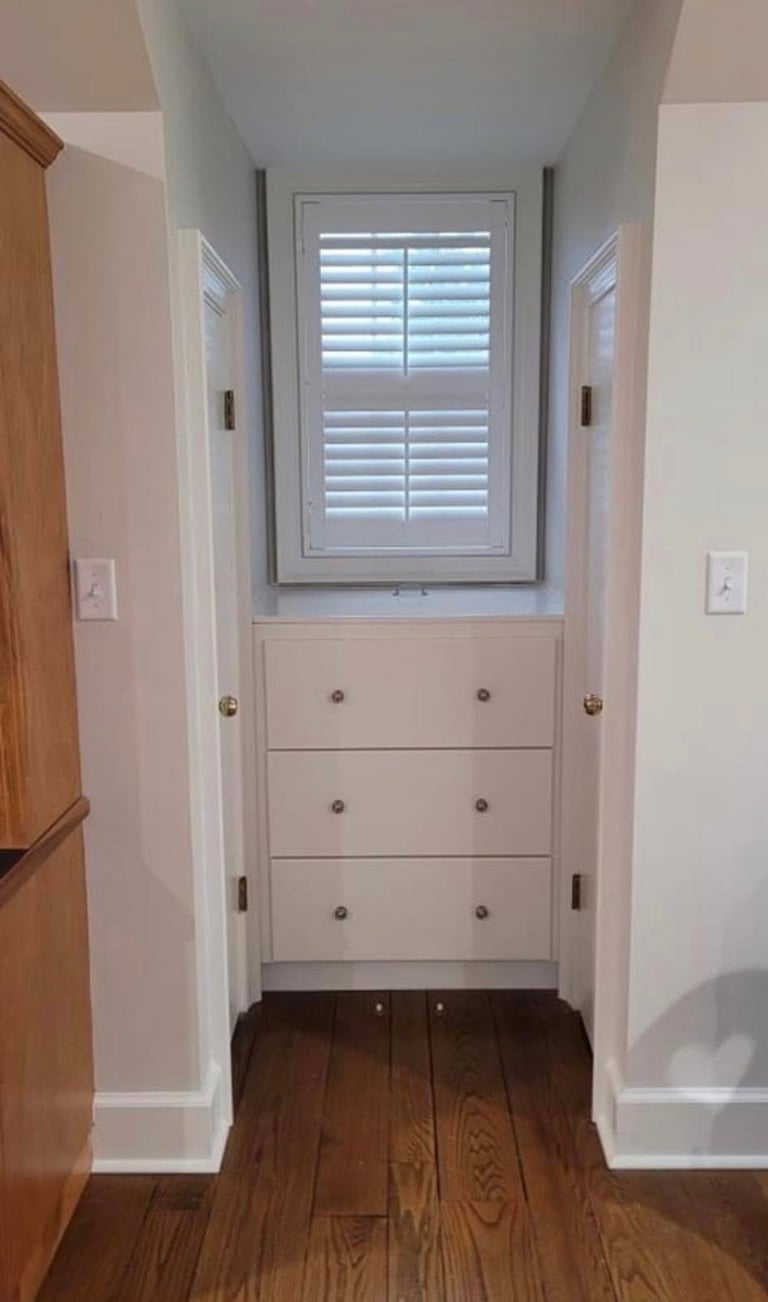 Hallway with white cabinetry and shuttered window above drawers, flanked by white walls and dark hardwood flooring