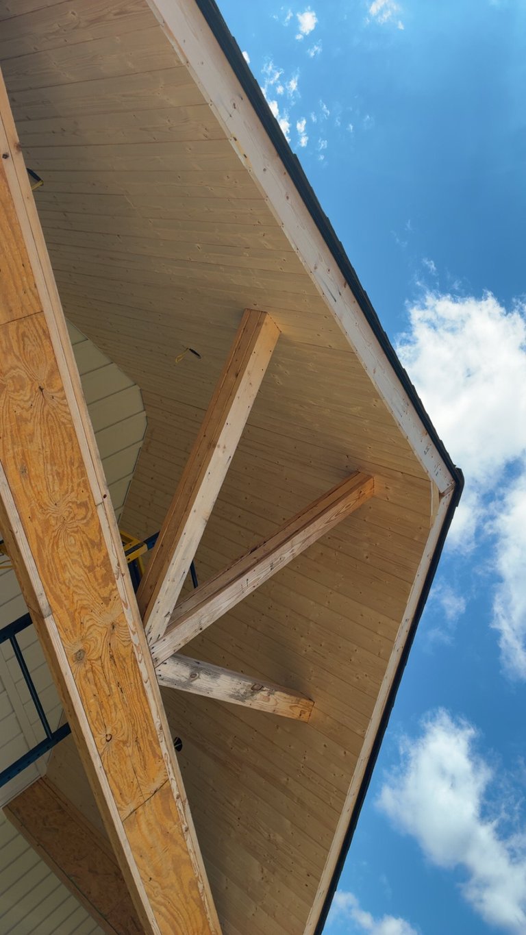 Upward view of a wooden house eave under construction with exposed beams and framing against a blue sky with clouds