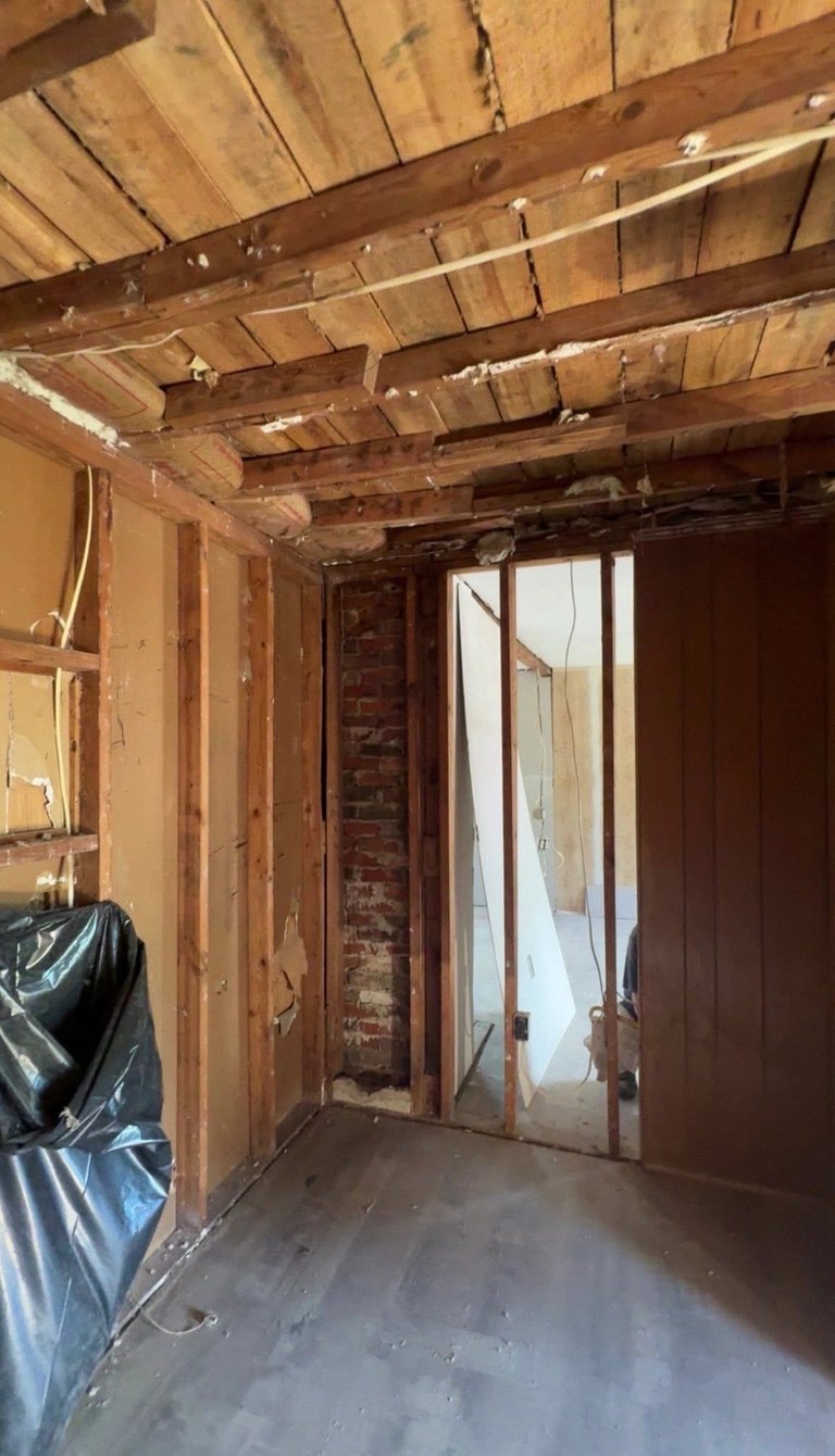 Interior of a gutted room under renovation with exposed wooden beams, brick walls, and plastic sheeting